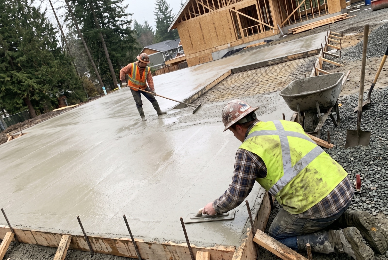 Two workers in hi-vis vests placing and finishing a concrete slab beside a house under construction